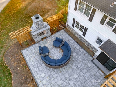 Top-down view of the new North Wilmington back patio: a curved modular wicker sectional with deep navy cushions wraps around the seating zone on the gray paver patio, the stand-alone fieldstone fireplace and cedar lattice screens visible at top of frame, and a strip of fresh black mulch borders the patio's edge