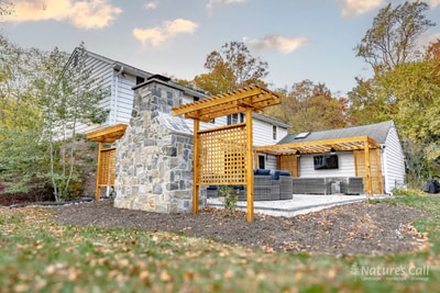 Wide ground-level view at peak autumn: the home's existing chimney rises at center, with the cedar pergola and lattice privacy screen at left, a second cedar pergola against the home at right, and the gray paver patio between them