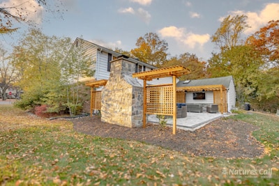 Side-angle ground view of the rebuilt North Wilmington back yard: the home's chimney rises at center, the cedar pergola and lattice privacy screen at left, and the curved wicker sectional and cedar pergola against the home visible to the right, all in fall light