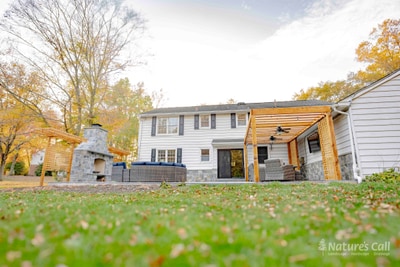 Wide ground-level view of the back of the home from across the lawn: the cedar pergola against the home shades the patio's seating zone, the stand-alone fieldstone fireplace stands at left, and a layer of fall leaves and a mature tree fill the foreground