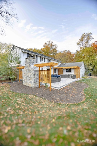 Mid-wide view of the patio's fireplace zone: the fieldstone outdoor fireplace anchors the left, cedar lattice screens flank both sides for privacy, and the gray paver patio and curved wicker sectional are in the foreground with the home's stone-clad rear wall behind