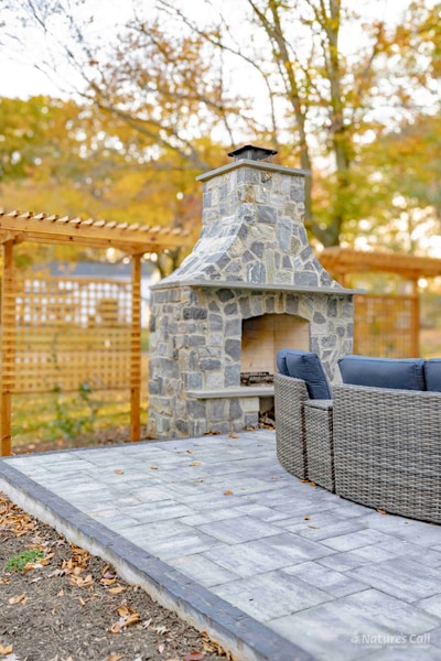 Close view of the stand-alone fieldstone outdoor fireplace with a stainless cooking grate and stacked firewood at the base, the curved wicker sectional with deep navy cushions in the right foreground, and the cedar pergola visible at upper left