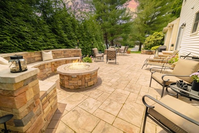 Stone fire pit lit at dusk in the center of a travertine paver patio, ringed by a curved natural-stone bench with cushions, with a dining set and lounge chairs beyond and a tall evergreen hedge enclosing the patio. Nature's Call Landscaping hardscaping project.