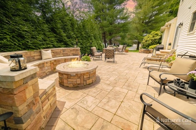 Stone fire pit lit at dusk in the center of a travertine paver patio, ringed by a curved natural-stone bench with cushions, with a dining set and lounge chairs beyond and a tall evergreen hedge enclosing the patio. Nature's Call Landscaping hardscaping project.