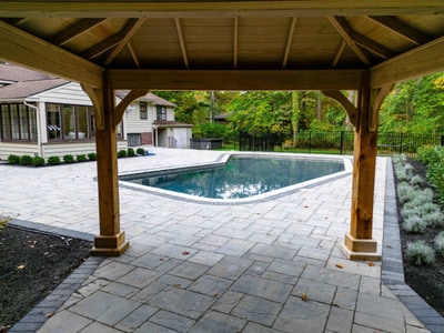 View from under a timber pavilion across a large gray paver pool deck to an in-ground pool, with a wooded backyard and aluminum fence beyond. Nature's Call Landscaping hardscape and pool deck project.