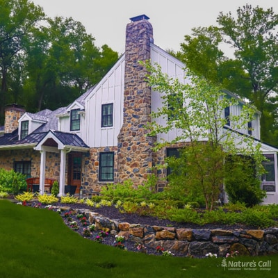 A white board-and-batten farmhouse with a fieldstone chimney, framed by layered foundation plantings and a natural-stone retaining wall with flowering beds along a curved lawn. Nature's Call Landscaping landscape design project.