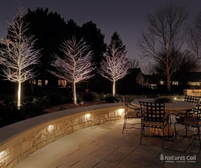 Three bare trees uplit against a deep blue night sky behind a curved natural-stone seat wall with recessed under-cap lighting, fronting a lit bluestone patio with a bistro table and chairs. Nature's Call Landscaping outdoor lighting project.