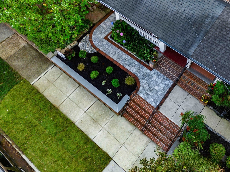 Drone view straight down on the Wilmington front entry: the geometric pattern of blue-gray pavers, red brick risers and edges, and the bluestone-capped planter bed read like a plan-view diagram of the hardscape