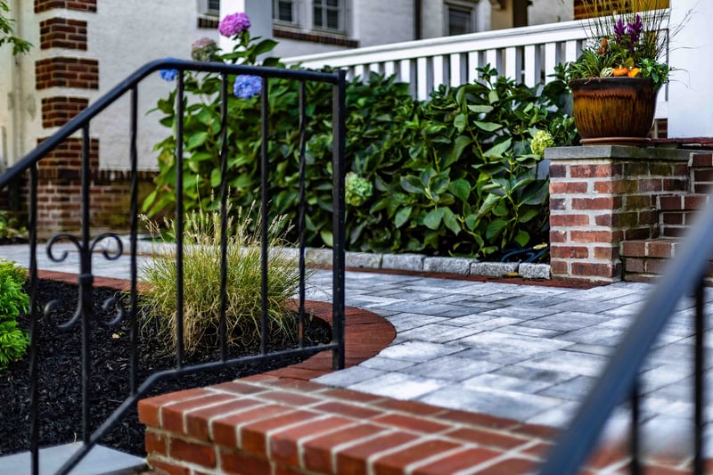 Magazine-style ground-level composition at the Wilmington paver landing: the scrollwork wrought iron railing on the left, an ornamental grass clump at center, the hydrangea hedge stretching back, and a brick column topped with a seasonal-flower urn at the right