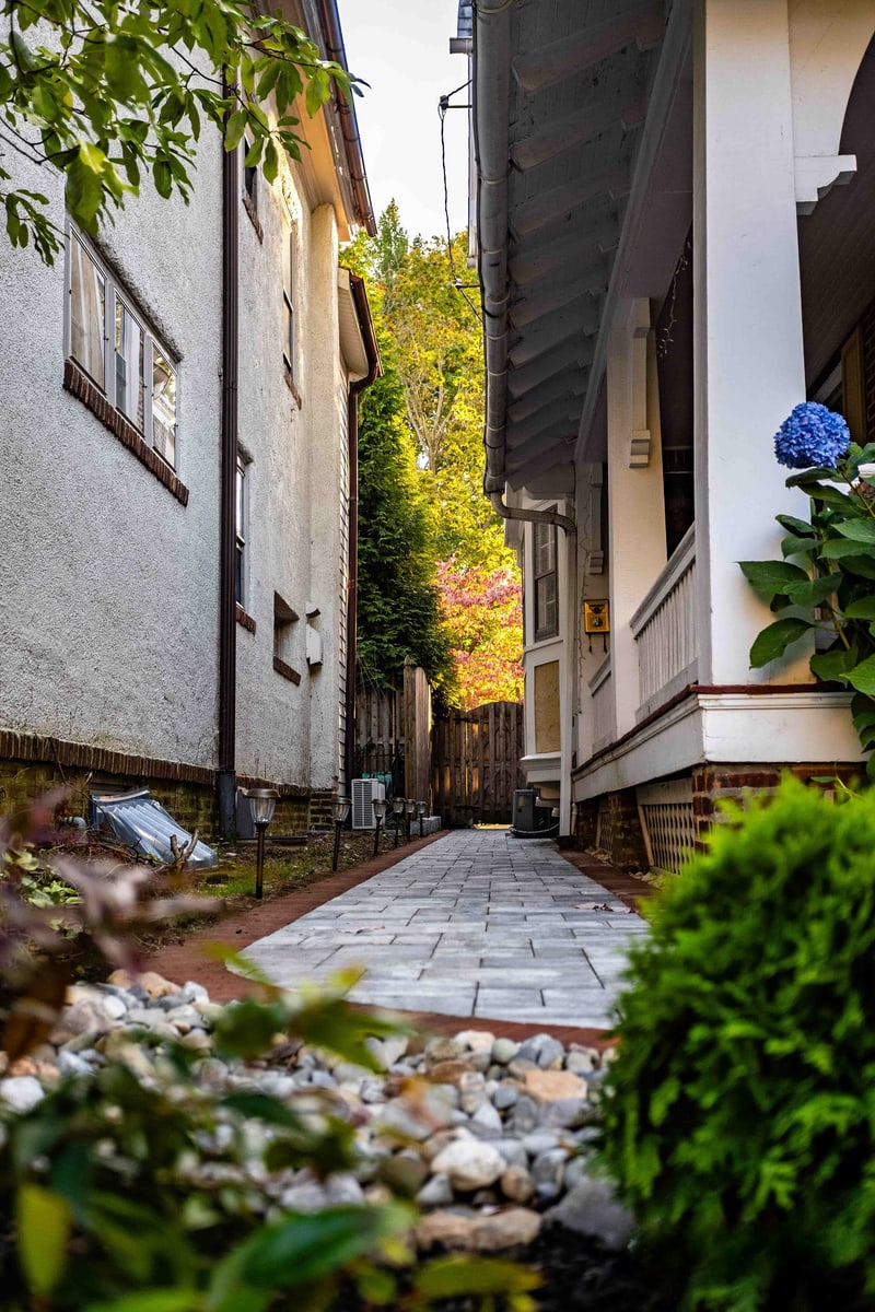 Cinematic vertical view of the side-yard paver alley running between two Wilmington homes: blue-gray pavers with red brick edge stretch toward an autumn-color tree at the alley's end, with a river-rock drainage strip and blue hydrangea bloom flanking the foreground