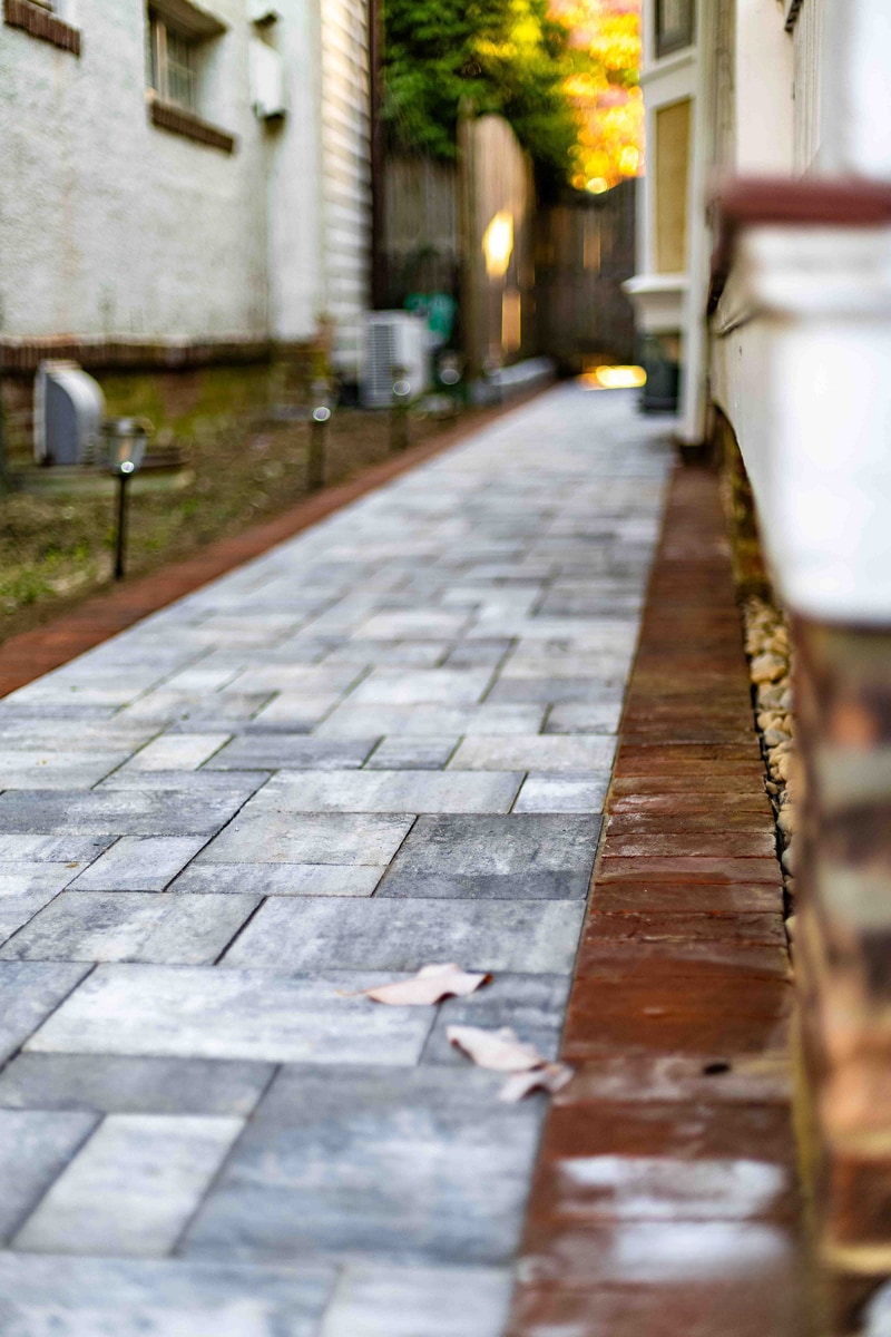 Cinematic vertical view down the Wilmington side-yard paver alley at golden hour: blue-gray pavers with red brick edge run to a glowing sunset spilling between the two homes, a path light at left and downspout at right anchoring the foreground