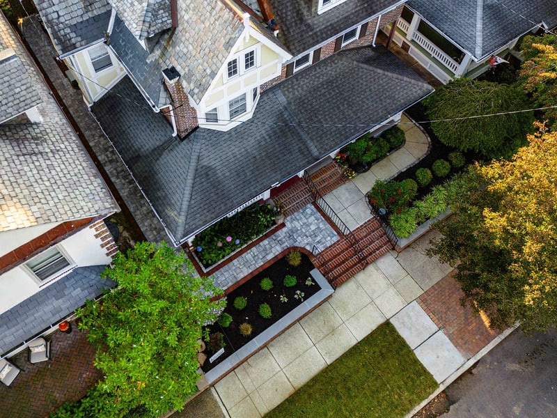 Aerial three-quarter view of the full Wilmington property: the cream-stucco Tudor-style home with gabled roof at center, the rebuilt front-yard hardscape laid out below, and the paver alley running along the right side