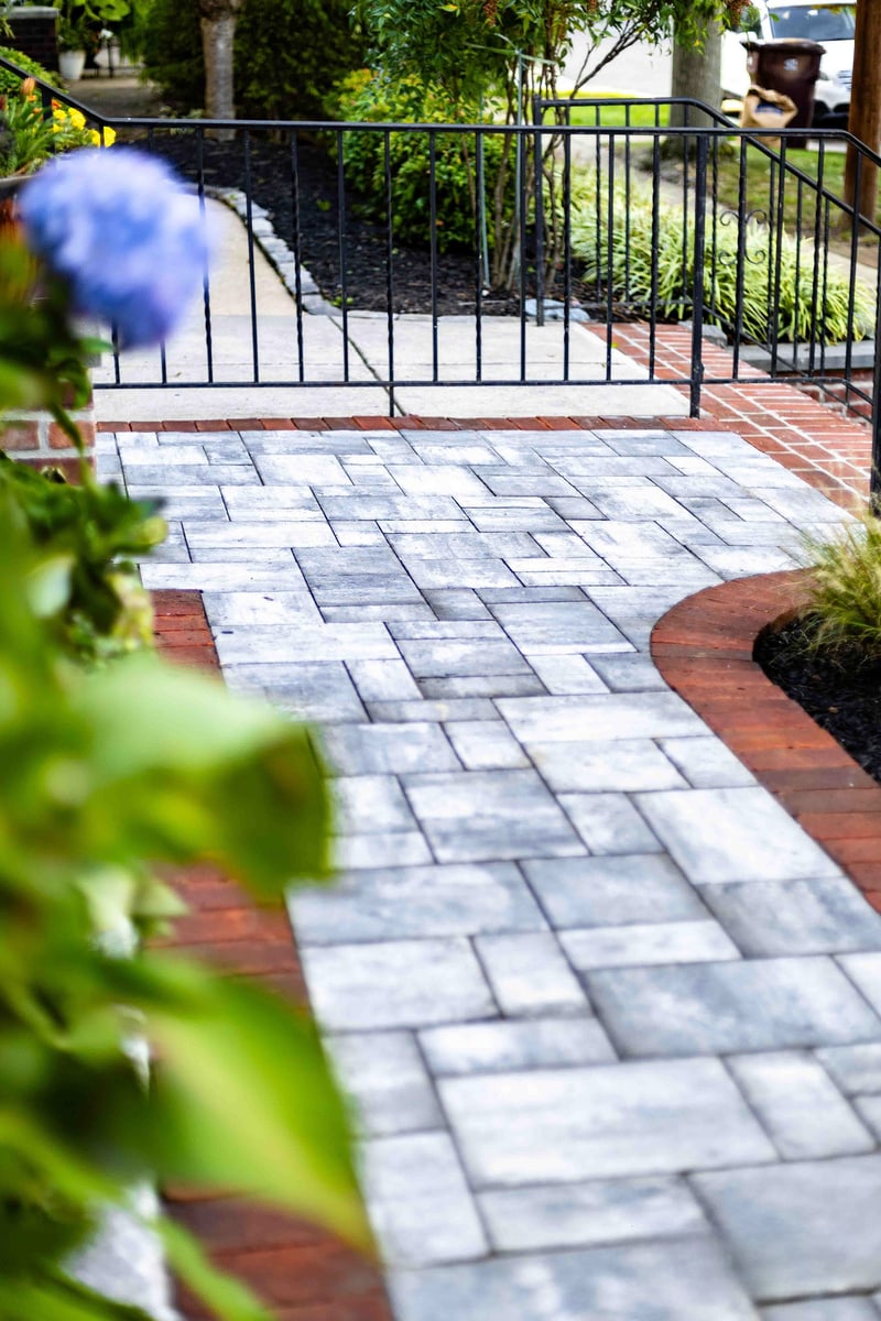 Vertical view down the curved paver walkway at the Wilmington front entry: blue-gray pavers with red brick edge sweep through the frame, a blue hydrangea bloom soft-focus in the foreground, scrollwork wrought iron railing and brick column visible at the path's end