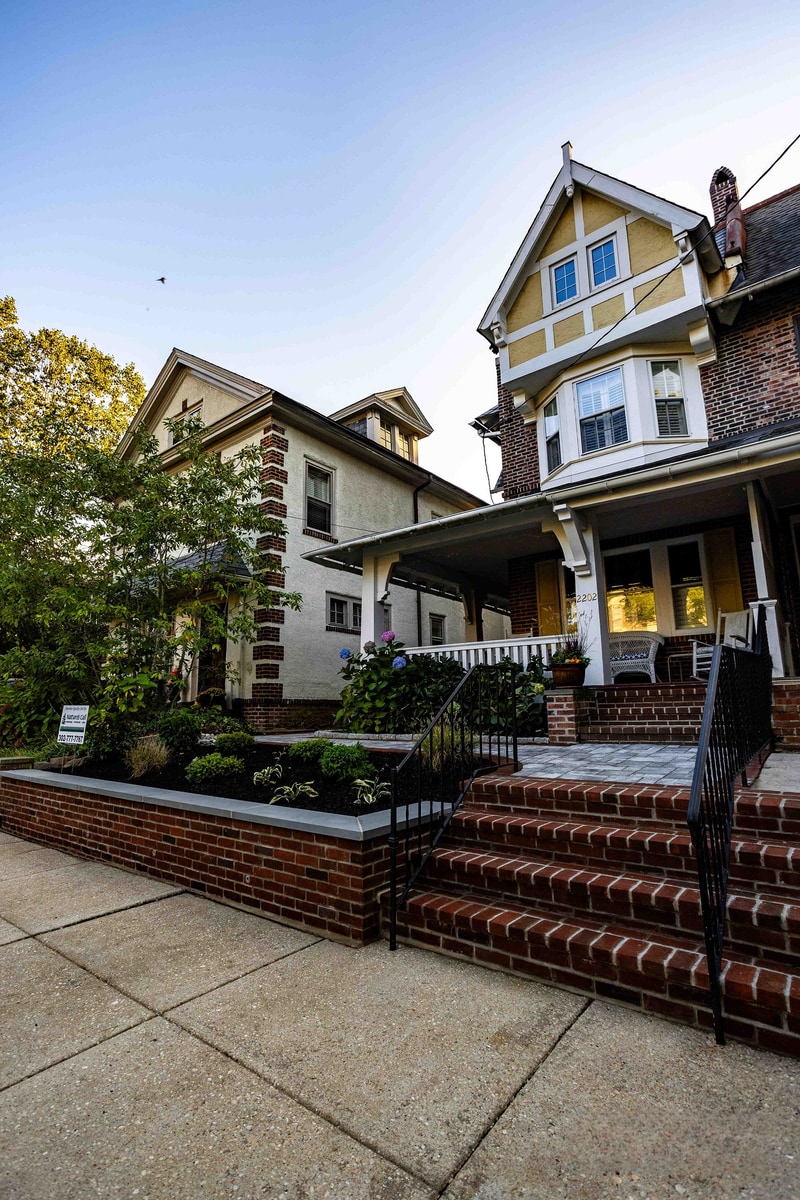 Wide street-level establishing view of the Wilmington property in context with neighboring homes: the cream-stucco Tudor-style facade with red brick accents and white trim, the rebuilt brick-faced planter wall along the sidewalk, and the porch entry visible past the hydrangea hedge