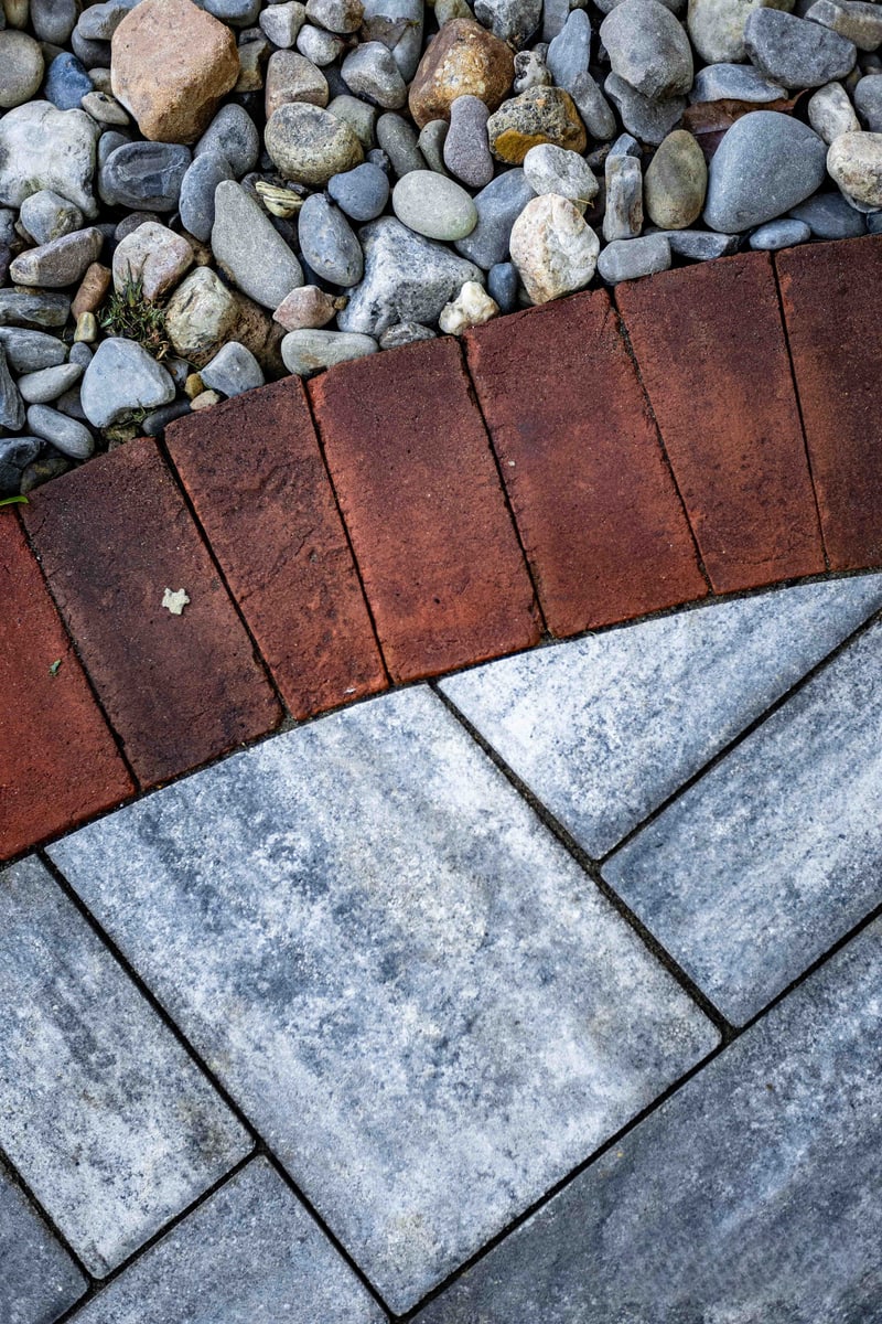 Vertical material macro at the Wilmington alley edge: blue-gray pavers in the foreground meeting the red brick curb, with a strip of river-rock drainage stones beyond — three of the project's core hardscape materials in one frame