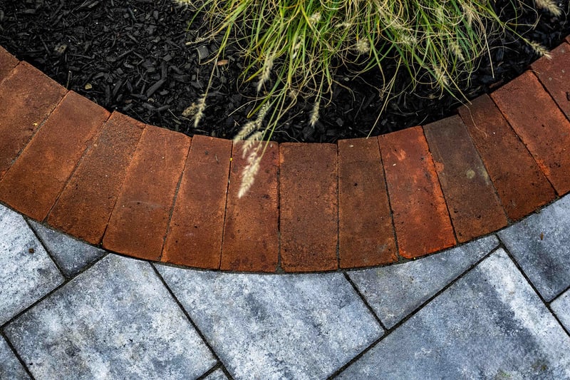 Close-up of the curved red brick edge meeting the blue-gray paver walkway in Wilmington, with an ornamental grass clump catching the light in the planter bed above the brick