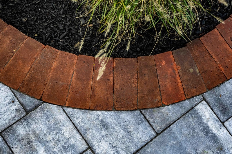 Close-up of the curved red brick edge meeting the blue-gray paver walkway in Wilmington, with an ornamental grass clump catching the light in the planter bed above the brick