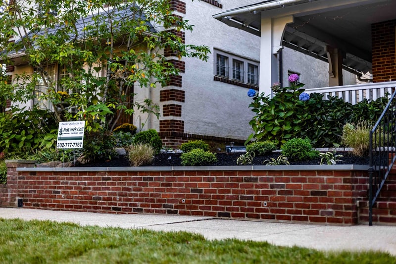 Side-street view of the Wilmington property showing the rebuilt brick-faced planter wall with bluestone cap running along the sidewalk, the Nature's Call yard sign at left, ornamental grasses and hostas in the bed, and a neighbor's home in the distance