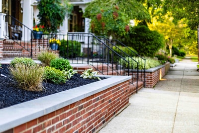 Side-on ground-level view of the Wilmington brick planter wall meeting the porch steps: ornamental grasses and hostas in the bed, scrollwork wrought iron railings on the steps, and the bluestone walkway running away from the camera