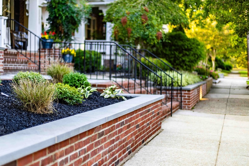 Side-on ground-level view of the Wilmington brick planter wall meeting the porch steps: ornamental grasses and hostas in the bed, scrollwork wrought iron railings on the steps, and the bluestone walkway running away from the camera