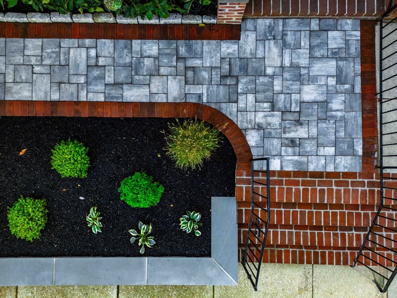 Drone view straight down on a Wilmington planting bed corner: the geometric meeting of blue-gray pavers, the curved red brick edge, and the mulch bed with mounded boxwoods, hostas, and a sweep of ornamental grass