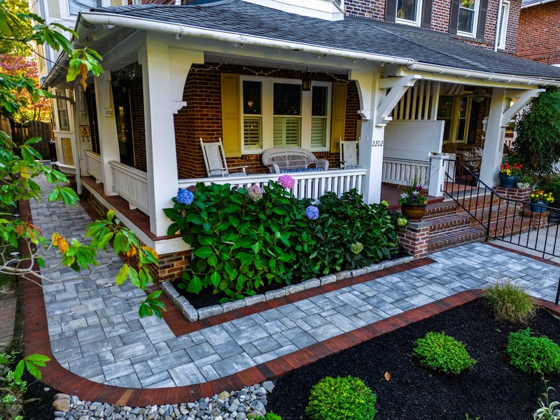 Front-yard view of a Wilmington Delaware row home with rebuilt front entry: a curved blue-gray paver walkway with red brick edging leads to a covered front porch, a hedge of blue and purple hydrangeas anchors the planter beds in the center, and brick-faced raised planters with bluestone caps frame both sides