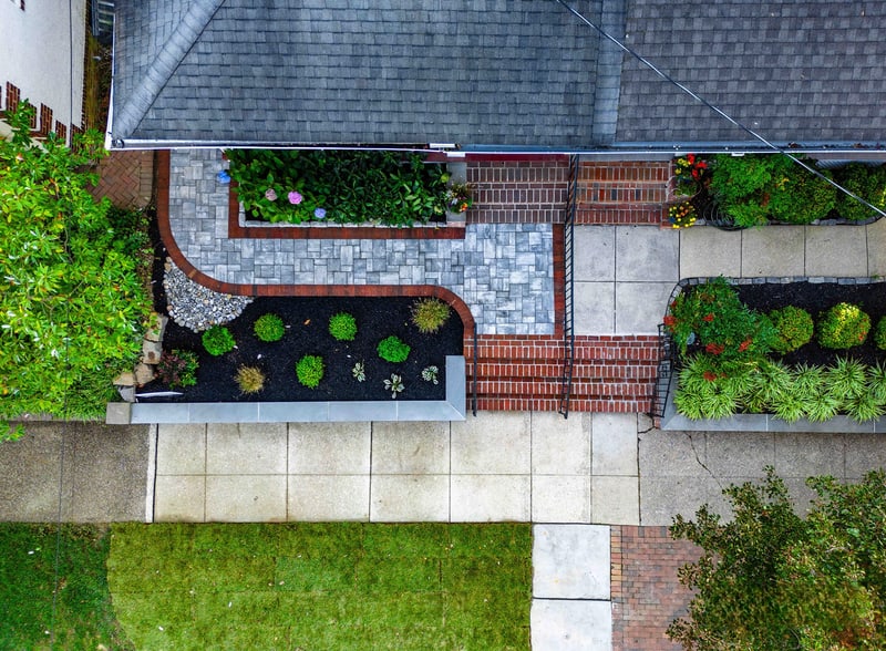 Drone view straight down on the Wilmington front yard: the curved paver walkway and brick-edged planter beds form a geometric pattern below the home's gable roofline, with mounded boxwoods and ornamental grasses in the mulch beds