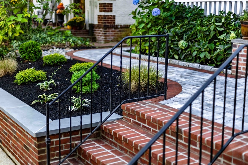 Ground-level view of the Wilmington paver landing detail: red brick risers in the foreground, scrollwork wrought iron railing curving up at left, planter bed with hostas and ornamental grasses to the side, and blue hydrangea hedge along the porch