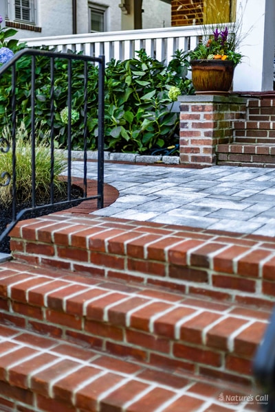 Portrait detail of the Wilmington brick stair risers leading up to the paver landing, with scrollwork wrought iron railing on the left, hydrangea hedge and a brick column with seasonal-flower urn at the top of the steps