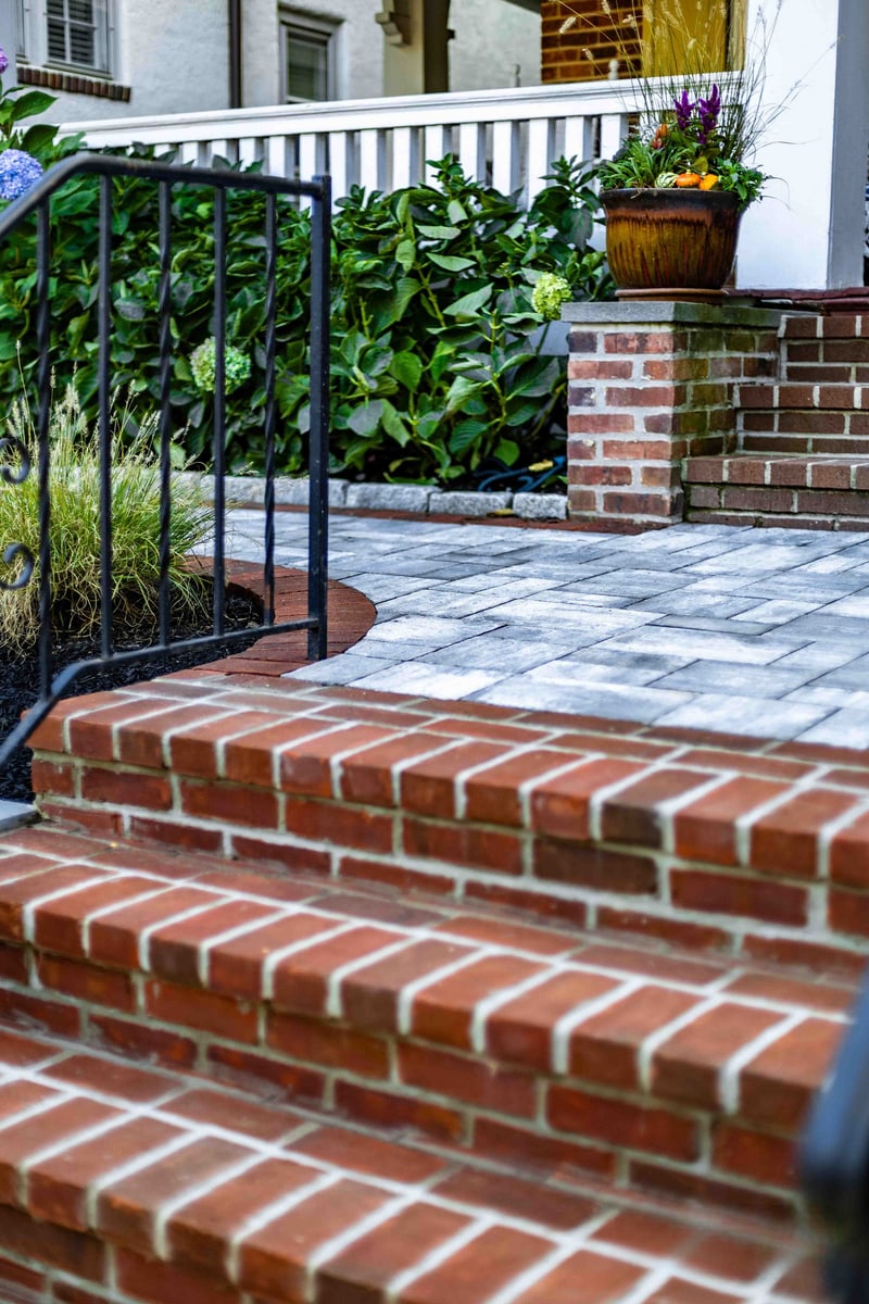 Portrait detail of the Wilmington brick stair risers leading up to the paver landing, with scrollwork wrought iron railing on the left, hydrangea hedge and a brick column with seasonal-flower urn at the top of the steps