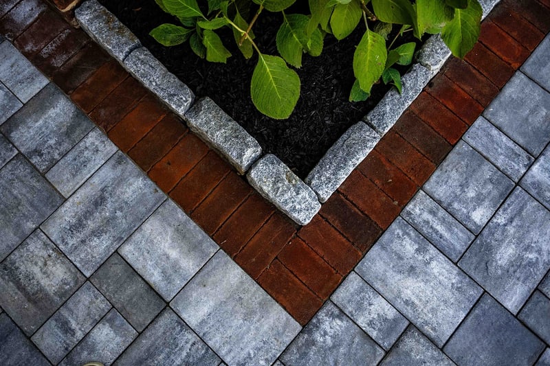Aerial close-up of the Wilmington paver-and-brick edge meeting at a corner: blue-gray pavers, red brick risers, granite cobble accent, and a hydrangea leaf reaching down from the planter above