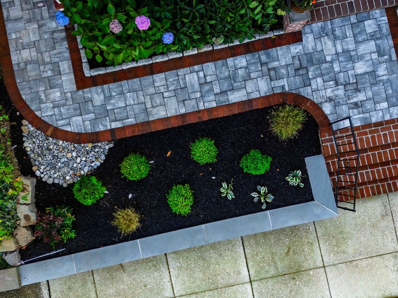Aerial view of the Wilmington planter bed corner: the curved red brick edge sweeps from the paver walkway around a mulched bed of mounded boxwoods, ornamental grass, and a small river-rock drainage well at the corner